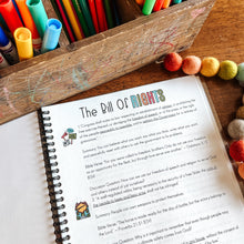Load image into Gallery viewer, Colorful markers and pens in a wooden box with an open Bill of Rights notebook on a wooden surface.