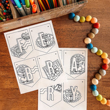 Load image into Gallery viewer, Coloring pages for February banner with stamp designs on a wooden table with coloring supplies.