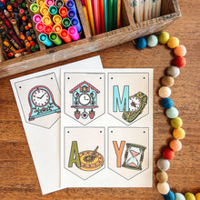 Load image into Gallery viewer, Colorful stationery items including crayons, markers, and colored pencils on a wooden surface with May banner.