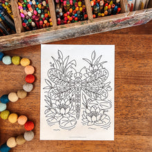 Load image into Gallery viewer, A black and white dragonfly coloring page with floral details, displayed on a wooden table surrounded by craft supplies including a box of colored pencils, markers, and felt beads.