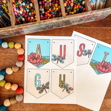 Load image into Gallery viewer, A dragonfly themed August banner arranged on a wooden table with an envelope, surrounded by craft supplies including a box of colored pencils and felt beads.