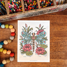 Load image into Gallery viewer, An art print featuring a colorful dragonfly and lotus flower design, displayed on a wooden table surrounded by craft supplies including colored pencils, markers, and pom-poms.
