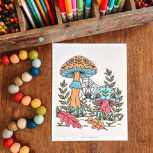Load image into Gallery viewer, A coloring page featuring two colorful mushrooms in a forest scene, displayed on a wooden table surrounded by art supplies, including markers, crayons, and painted beads.
