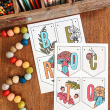 Load image into Gallery viewer, October banner with autumn-themed illustrations are arranged on a wooden table, surrounded by colorful craft supplies including markers, pencils, beads, and paintbrushes.