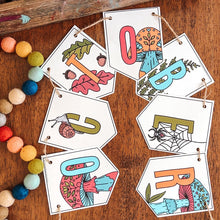 Load image into Gallery viewer, A decorative pennant banner spelling October hangs on a wooden table, with colorful art supplies and multicolored beads nearby. The letters are illustrated with autumn-themed designs like leaves, acorns, spiders, and fall foliage.