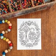 Load image into Gallery viewer, A black-and-white line art coloring page featuring a decorative key and heart design, placed on a wooden tabletop with colorful art supplies around it, including a divided tray of crayons and a strand of painted beads.