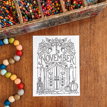 Load image into Gallery viewer, A black and white coloring page featuring a fall door design with the word 'November' and pumpkins, placed on a wooden tabletop next to a wooden pencil box and colorful beads.