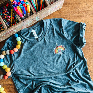 dark grey t-shirt with a rainbow and arrow design on a wooden surface with art supplies. Psalm 127:4