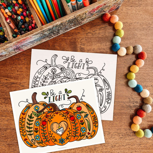 Coloring books with pumpkins and text 'Be a Light' on a wooden surface with coloring supplies.
