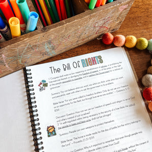 Colorful markers and pens in a wooden box with an open Bill of Rights notebook on a wooden surface.