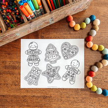 Load image into Gallery viewer, Coloring page with gingerbread-themed shapes on a wooden surface with art supplies.
