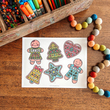 Load image into Gallery viewer, Coloring sheet with decorated gingerbread people on a wooden surface with coloring supplies.
