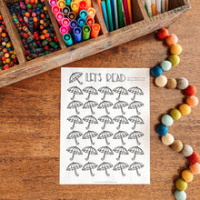 Load image into Gallery viewer, Colorful markers, a reading tracker coloring sheet with umbrellas, and decorative items on a wooden surface.
