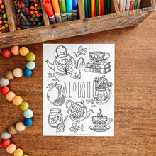 Load image into Gallery viewer, Coloring sheet with 'APRIL' and tea-themed illustrations on a wooden surface with art supplies.
