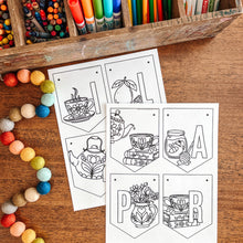 Load image into Gallery viewer, Coloring page April banner with tea-themed designs on a wooden surface with art supplies.
