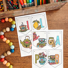 Load image into Gallery viewer, Colorful stationery items including crayons, markers, and colored pencils in a wooden box on a wooden surface with tea themed April banner.
