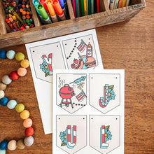 Load image into Gallery viewer, Colorful stationery items including markers, pens, and a wooden box on a wooden surface with July banner coloring sheets.
