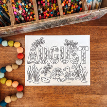 Load image into Gallery viewer, A white coloring card featuring the word 'AUGUST' in decorative outlined letters with pond, dragonfly, and duck illustrations, placed on a wooden tabletop. A rustic box of colorful crayons and a string of multicolored pom-poms are visible around it.
