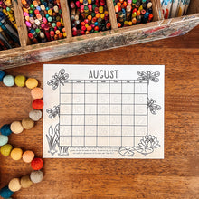 Load image into Gallery viewer, A printable August coloring calendar sheet with dragonfly and floral illustrations, placed on a wooden table surrounded by colorful crayons and felt beads.
