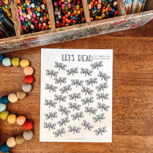 Load image into Gallery viewer, A children's coloring page featuring the text 'LET'S READ' and rows of dragonfly illustrations, styled on a wooden table with colorful art supplies and felt beads.
