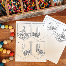 Load image into Gallery viewer, Black and white printable August banner arranged on a wooden tabletop. A wooden tray of colorful pencils and crayons sits in the background, and multicolored pom-poms line the left side.
