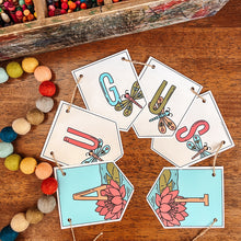 Load image into Gallery viewer, A colorful August banner with decorative dragonfly and flower illustrations, arranged on a wooden surface with a pencil case and colorful pom-poms nearby.
