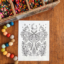 Load image into Gallery viewer, A black-and-white coloring page featuring a detailed moth and floral design, styled on a wooden tabletop with a wooden pencil tray, colorful pom-poms, and crayons nearby.
