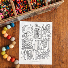Load image into Gallery viewer, A black and white printable coloring page with the word 'SEPTEMBER' in decorative lettering and floral/butterfly illustrations, placed on a wooden table with a box of colored pencils and pastel beads nearby.
