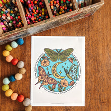 Load image into Gallery viewer, An art print featuring a colorful illustration of moths, a crescent moon, flowers, and stars, styled on a wooden table with a wooden pencil tray and colorful beads nearby.
