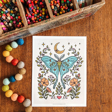 Load image into Gallery viewer, A coloring page featuring a detailed illustration of a blue-green luna moth surrounded by flowers, leaves, and a crescent moon, displayed on a wooden table next to a wooden pencil box and colorful beads.
