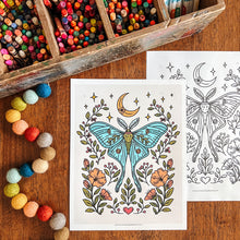 Load image into Gallery viewer, A coloring page set displayed on a wooden table, with one page showing a colored illustration of a luna moth surrounded by flowers, and another page showing the black-and-white line art. A wooden tray filled with colored pencils and a string of colorful pom-poms are arranged around it.

