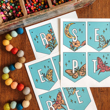 Load image into Gallery viewer, Printed September banner flags laid out on a wooden table next to a wooden box of colorful pencils and a string of multicolored pom-poms. The flags feature a light blue background with white letters for the months and illustrations of flowers.
