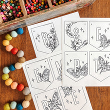 Load image into Gallery viewer, Black and white printable coloring banner with moths and floral designs arranged on a wooden table next to a wooden box of colored pencils and a string of colorful pom-poms.
