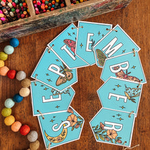 Load image into Gallery viewer, A set of teal monthly banner flags with white lettering and colorful insect and floral illustrations, arranged in a semi-circle on a wooden tabletop. A wooden box of colored pencils and a string of colorful beads are visible in the background and left foreground.
