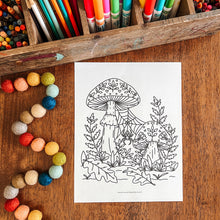 Load image into Gallery viewer, A black and white line art coloring page of a mushroom forest, displayed on a wooden table surrounded by colorful crayons, markers, and felt beads.
