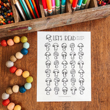 Load image into Gallery viewer, A children's coloring page featuring black mushroom illustrations and the text 'LET'S READ' on a white sheet, styled on a wooden table with crayons, colored pencils, and colorful beads nearby.
