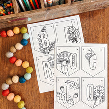 Load image into Gallery viewer, Two printable black-and-white lettering cards laid on a wooden table, surrounded by colorful craft supplies including markers, paintbrushes, beads, and crayons. The cards show decorative letter outlines for autumn-themed spelling.
