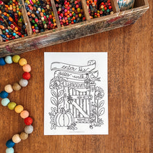 Load image into Gallery viewer, A black and white coloring page featuring a fall gate illustration with the text 'Enter the Gates of Thanksgiving' and 'LARKSSWIT', placed on a wooden tabletop surrounded by colorful crayons, beads, and a wooden pencil box.
