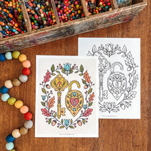 Load image into Gallery viewer, Two printable coloring pages with ornate key and padlock illustrations are arranged on a wooden surface, with one page colored in and one page uncolored. A wooden tray of colorful pencils and a string of multicolored beads are visible around them.
