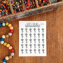 Load image into Gallery viewer, A printable reading tracker page titled 'LET'S READ' with rows of black key illustrations on a white sheet, styled on a wooden tabletop with a box of colorful pencils and multicolored beads nearby.
