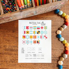Load image into Gallery viewer, Sheet of paper with nautical flag symbols on a wooden surface with colorful markers and pom-poms.
