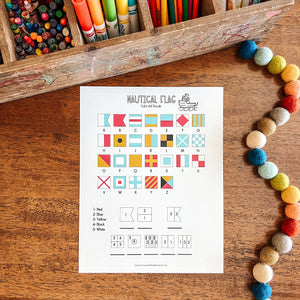 Sheet of paper with nautical flag symbols on a wooden surface with colorful markers and pom-poms.